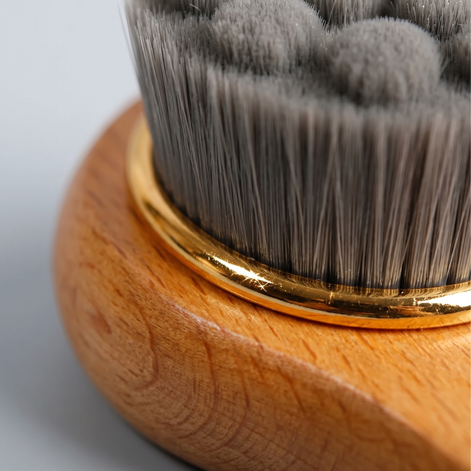 Close-up of a makeup brush with gray bristles, a shiny gold metal ferrule, and a round light brown wooden handle on a light gray background.