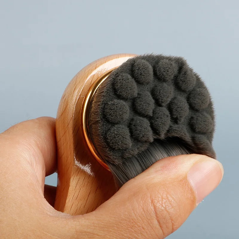 Hand holding a wooden facial cleansing brush with a gray bristle head against a light gray background
